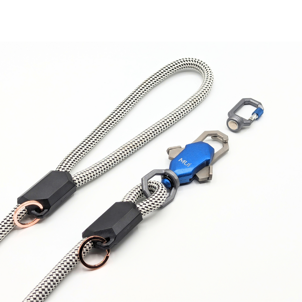 A close-up of a braided silver and black strap with black plastic connectors, metal rings, and a blue anodized metal clip labeled 'M6'. A separate matching clip is shown beside it on a clean white background.